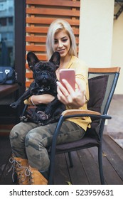 Beautiful and happy blonde woman enjoying in cafe bar and taking selfie photo with her adorable French bulldog. Selective focus on woman.