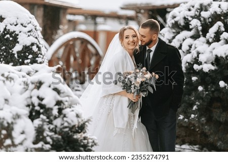 Beautiful and happy bride and groom are having fun among snowy trees. Bride and groom in the winter park. Bride with a bouquet of flowers in a wedding dress and poncho. Groom in a black coat.