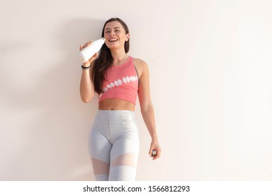 Beautiful Happy Brunette Fitness Model And Wearing A Pink Crop Top And Gray Yoga Tights Standing Against A Plain Beige Wall With A White Water Bottle