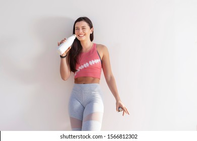 Beautiful Happy Brunette Fitness Model And Wearing A Pink Crop Top And Gray Yoga Tights Standing Against A Plain Beige Wall With A White Water Bottle