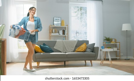 Beautiful And Happy Brunette Young Woman Moving Towards The Washing Machine In Homely Jeans Clothes. She Carrying Laundry Basket To The Washer. Bright And Spacious Living Room With Modern Interior.