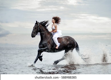 Beautiful happy caucasian woman in white flying dress riding without saddle on glorious shiny black frisian horse galloping by sea water making big nice splashes on surface 