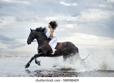 Beautiful happy caucasian woman in white dress riding without saddle on glorious shiny black frisian horse galloping by sea water making big nice splashes on surface