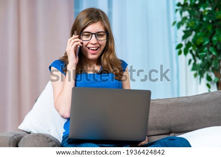 Beautiful happy cheerful girl, young excited positive woman in glasses is looking at screen of her laptop computer and calling, talking on cell mobile phone at home on sofa or couch in living room.