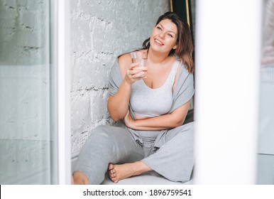 Beautiful happy emotional brunette young woman plus size body positive in comfortable sport wear with glass of water at home