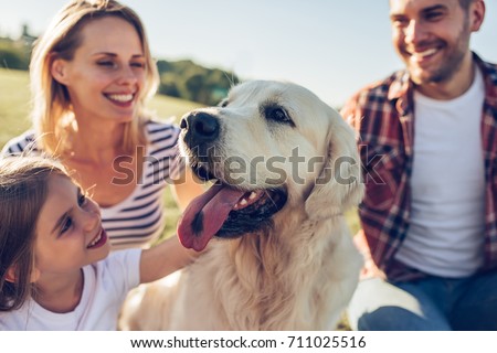 Beautiful happy family is having fun with golden retriever outdoors. Mother, father and daughter are sitting with dog labrador on green grass in park.