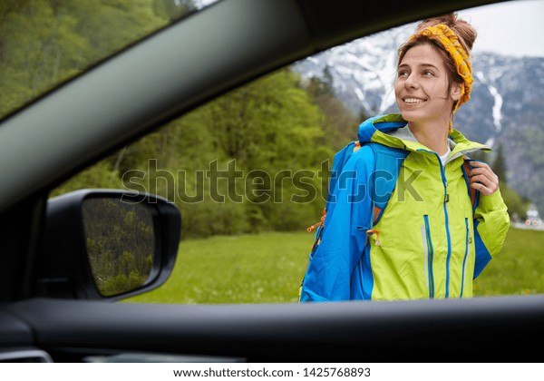 Beautiful happy female explorer poses against high\
mountains and green forest, somebody photographs traveler from car.\
Cheerful tourist focused away, enjoys views. Picturesque nature\
area around