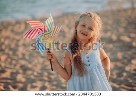 beautiful happy girl 6-7 years old having fun on sea sandy beach. vacation with children, trips and travel with children, preschool child smiling, playing with a toy windmill. holidays, summer camp