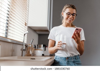 Beautiful Happy Girl In Eyeglasses Drinking Water While Using Mobile Phone At Home Kitchen