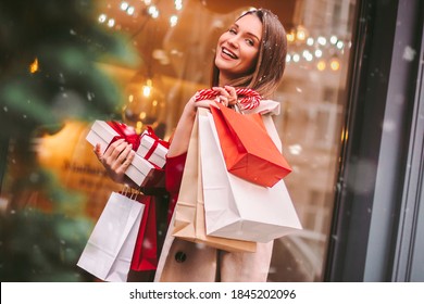 Beautiful Happy Girl Hold Shopping Bags And Gift Boxes In Hands And Smile On City Street. Young Attractive Cheerful Woman With Paper Bags And Present Box After Christmas Shopping In Mall. Winter Sale