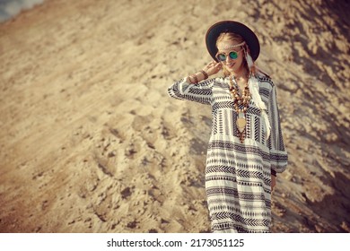 A beautiful happy girl in a loose ethnic dress, hat and boho jewelry walks along the ridge of a sand dune and smiles. Boho and hippie style. Summer, vacation and travel. Copy space.