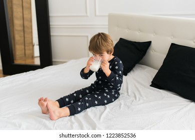 Beautiful Happy Kid Drinking Milk In The Morning While Sitting On The Bed Wearing One Piece Pajamas. Boy Drinking Glass Of Milk. 