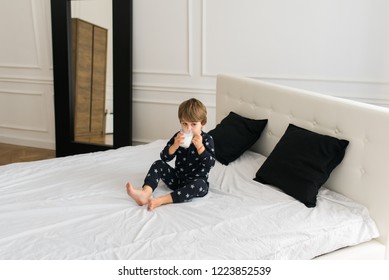 Beautiful Happy Kid Drinking Milk In The Morning While Sitting On The Bed Wearing One Piece Pajamas. Boy Drinking Glass Of Milk. 