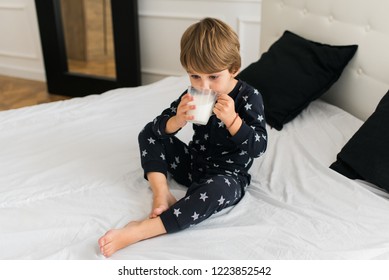 Beautiful Happy Kid Drinking Milk In The Morning While Sitting On The Bed Wearing One Piece Pajamas. Boy Drinking Glass Of Milk. 