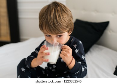 Beautiful Happy Kid Drinking Milk In The Morning While Sitting On The Bed Wearing One Piece Pajamas. Boy Drinking Glass Of Milk. 