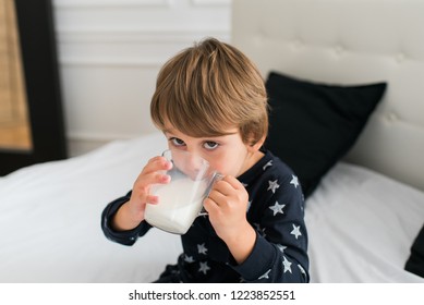 Beautiful Happy Kid Drinking Milk In The Morning While Sitting On The Bed Wearing One Piece Pajamas. Boy Drinking Glass Of Milk. 