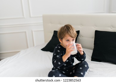 Beautiful Happy Kid Drinking Milk In The Morning While Sitting On The Bed Wearing One Piece Pajamas. Boy Drinking Glass Of Milk. 