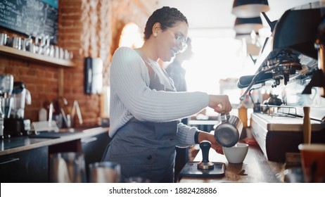 Beautiful and Happy Latin American Female Barista with Short Hair and Glasses is Making a Cup of Cappuccino in Coffee Shop Bar. Male Cashier Works at a Cozy Loft-Style Cafe Counter in the Background. - Powered by Shutterstock