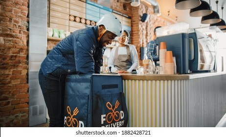 Beautiful Happy Latin Barista Serves Order To A Food Delivery Courier Picking Up Paper Bag With Pastries From A Cafe Restaurant. Delivery Guy Puts Food In His Hot Thermal Insulated Bag.