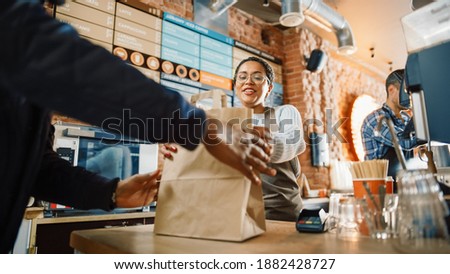 Beautiful Happy Latin Barista Serves Order to a Food Delivery Courier Picking Up Paper Bag with Pastries from a Cafe Restaurant. Delivery Guy Puts Food in His Hot Thermal Insulated Bag.