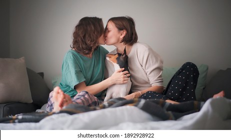 Beautiful happy lesbian couple in pajamas sitting on bed in the morning with pet dog bassengi tenderly kissing and hugging - Powered by Shutterstock