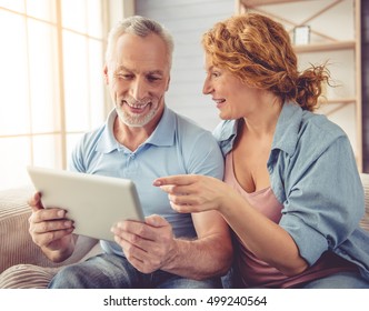 Beautiful Happy Mature Couple Is Using A Digital Tablet, Talking And Smiling While Sitting On Couch At Home