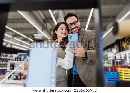 Beautiful and happy middle age couple buying consumer tech products in modern home appliances store. They are choosing smart phone or digital tablet. People and consumerism concept.
