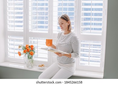 Beautiful happy middle age woman sitting on windowsill at home and reading book. Young woman with short hair drinking tea coffee from mug and holding reading book. Relax, leisure time at home. - Powered by Shutterstock