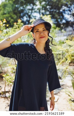 Beautiful happy middle-aged Japanese woman in black casual dress and straw sun brim hat in summer park, smiling. Sunlight, copy space