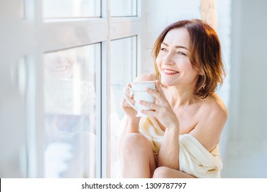 Beautiful Happy Miling Mature Woman With Clear Skin And Cute Smile. Female Sitting On Windowsill Drinking Tea. Elegant, Vulnerable, Fond, Gentle Concept. Spa And Skincare.