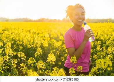 Beautiful Happy Mixed Race African American Girl Teenager Female Young Woman Athlete Runner Drinking Water From A Bottle In A Field Of Yellow Flowers At Sunset In Golden Evening Sunshine