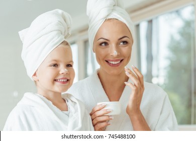 Beautiful Happy Mother And Daughter In Bathrobes And Towels Applying Face Cream Together