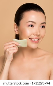 Beautiful Happy Nude Asian Woman Making Massage Of Face With Jade Gua Sha Stone Isolated On Beige