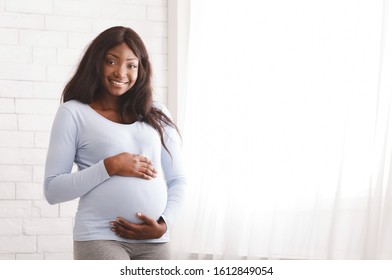 Beautiful happy pregnant black woman touching her tummy, staying next to window, posing at home, free space