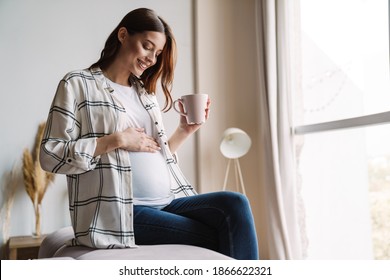 Beautiful happy pregnant woman smiling and drinking coffee while sitting on couch at home