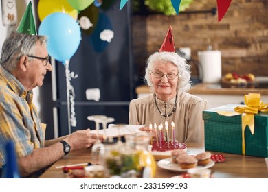 Beautiful happy senior couple sitting at kitchen table in birthday hats, elderly man giving a birthday present to her wife. Lifestyle, senior life, togetherness concept. - Powered by Shutterstock