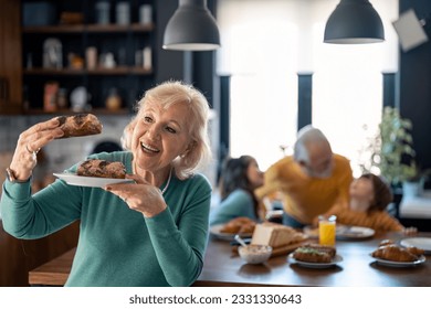 Beautiful happy senior woman in selective focus holding a plate and looking at sweet delicious pastries while standing in the kitchen with senior man and two kids playing at table in the background. - Powered by Shutterstock