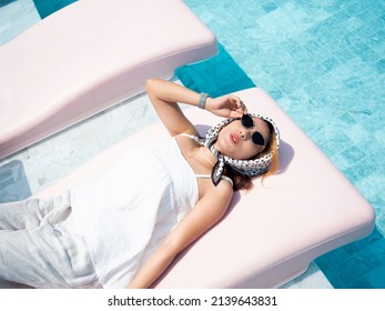 Beautiful Happy Smile Asian Woman Portrait In Casual White Shirt Wearing Sunglasses And Hair Scarf Reclining On Pink Sunbed Near The Pool, Enjoy The Vacation On Sunny Day In Summer.