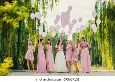 Beautiful happy smiling bride with bridesmaids in light trendy pink dresses on walk outdoors holding pink balloons in green summer city park. Friends, maid of honor, female friendship, wedding concept