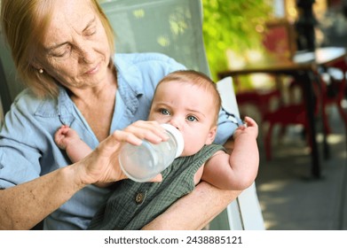 Beautiful happy smiling senior elderly woman nanny feeding formula on hands from bottle cute little baby boy. Grandmother and grandson at sidewalk cafe at summer day. Multigenerational family concept - Powered by Shutterstock