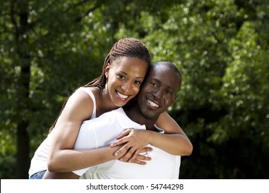 Beautiful Happy Smiling Young African American Couple In Love Wearing White Shirts, Woman Hugging Man, With Trees In The Background.