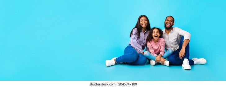 Beautiful And Happy Smiling Young Black Family Laughing And Having Fun Time Together While Sitting On The Floor And Looking At Camera, Isolated On Blue Studio Background, Free Copy Space, Panorama