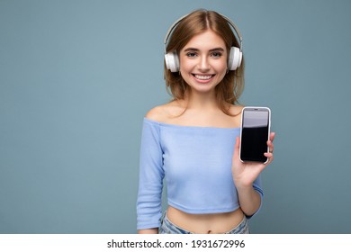 Beautiful happy smiling young woman wearing stylish casual outfit isolated on background wall holding and showing mobile phone with empty display for mockup wearing white bluetooth headphones