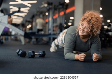 Beautiful happy smiling young woman in comfortable sports clothes feeling motivated determined to work on her body weight to stay active, healthy and in shape practicing plank exercise at gym. - Powered by Shutterstock