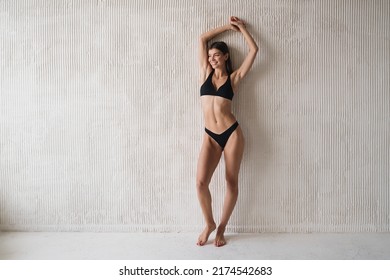 Beautiful Happy Woman In Black Fitness Bikini Posing With Raised Hands On White Textured Wall With Empty Space For Your Content. People And Sports. Healthy Lifestyle. Flat Stomach