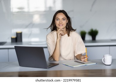 Beautiful happy woman drinking tea while working with laptop in cozy kitchen