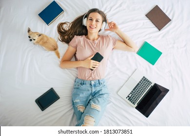 Beautiful happy woman in headphones is lying on the bed with her cute little dog and listening to music