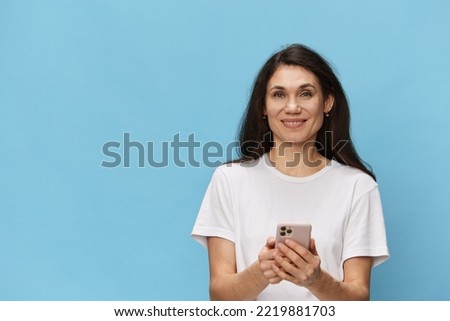 beautiful, happy woman in a light T-shirt stands holding a phone in her hands on a light blue background