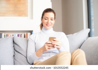 Beautiful Happy Woman With Mobile Phone Sitting On Sofa At Home And Using Mobile Banking. 