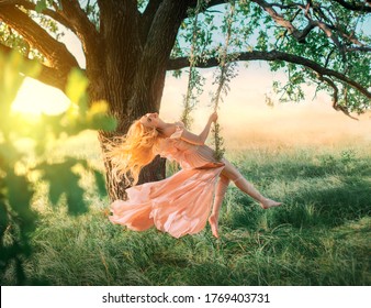 Beautiful happy woman nymph sitting on swings. Magical fantasy swing. princess long peach color orange silk vintage dress fluttering wind. blond hair fly in motion. Tree sunshine green grass forest